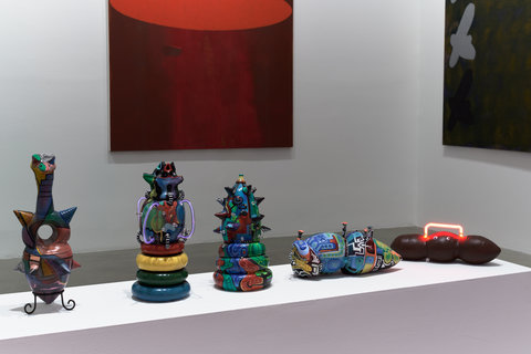 Installation view of the exhibition featuring a white bench shaped pedestal. The pedestal has five sculptures placed atop it in a row. The sculptures are multicolored and extremely abstract. There is a red painting hanging out of focus against a whitewall in the background. 