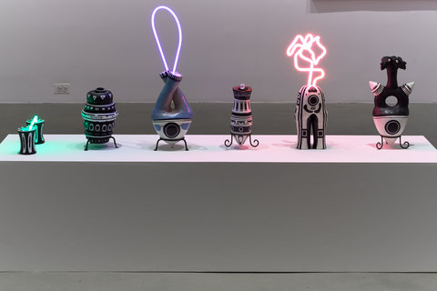 Image of six sculptures on a white bench in the exhibition. Some of the sculptures include neon light components of purples, pinks, and blues. 