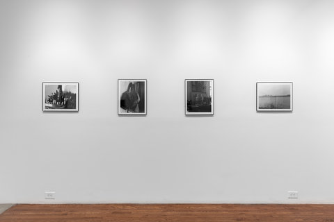 Installation view of four photographs in the exhibition. Four black and white photographs are hung evenly spaced on a white wall. The first image is dark, but features what looks like makeup in a organizer on a desk. The next photograph features a bag hanging on the the top corner of a door. The next photograph features a vase, or urn, on a table surrounded by various objects. And the final image, is a landscape of water with a distant city in the background. 