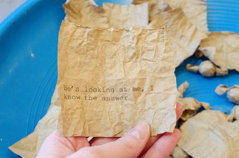 Close up image of a torn piece of brown paper in someones hand. In black type face, the paper states, "HE'S LOOKING AT ME, I KNOW THE ANSWER."