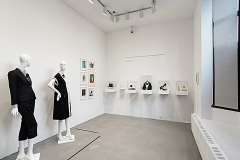 Two mannequins wearing black tuxedo-like dresses sit in a gallery with several framed images and displays.