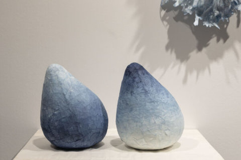 Image of two sculptures on a white pedestal in the exhibition. The close-up image features two tear drop shaped sculptures made of blue and white paper maches. 