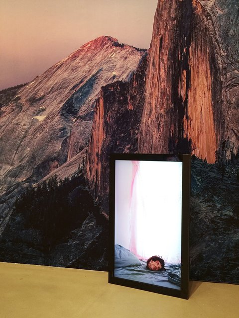 Image of an object in the exhibition featuring a monitor with a still of a severed head lying on a blue bed sheet. Behind the monitor a large mountain scene unfolds. 