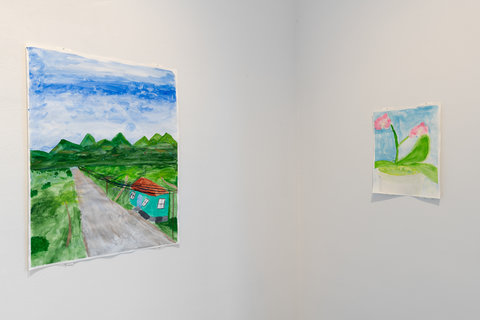 Installation view of two works in the exhibition. On the left wall is a grey road as it disappears into the distance. Alongside the road is a small turquoise house with a red-shilling roof. In the background there are blue skies and mountains. On the right wall is a painting of two pink flowers with green stems and leaves. The background is blue on the top and white on the bottom. 