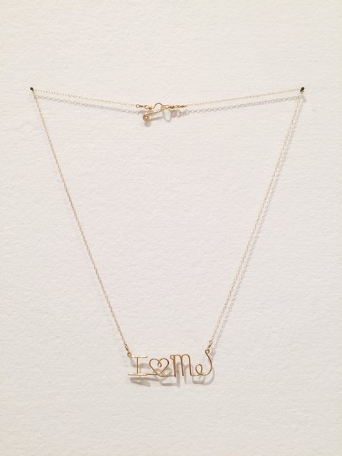 Close-up image of a sculpture from the exhibition featuring a necklace-like chain hanging on two nails on the wall. The gold chain hangs down to a charm that reads I HEART ME with a heart where the word heart would be. The writing is in loopy, cursive like letters.