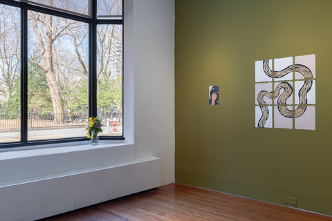 Installation view of the exhibition. A large window allows light to pour in from the left side of the image. On the right side, on an dark olive green colored wall, are two works of art. The first work is a drawing of a hand that seems to be touching shining black hair. Next to this work, is a set of nine drawings in three rows of three. A squiggly line cuts across all of the pages as if it were one work, split apart into nine pieces. The line is very thick and outlined in black and has light brown hatch marks in smaller branch patterns inside the outline. It is drawn on a white background. 