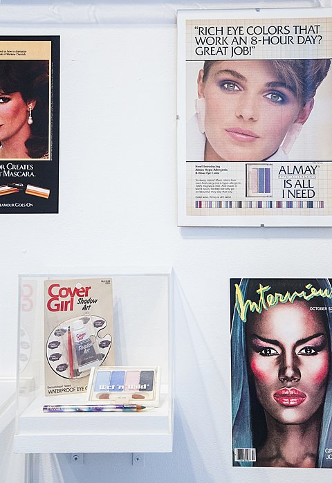 Framed prints of makeup advertisements hanging on a wall, along with a display case holding vintage eye shadow.