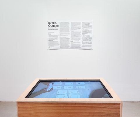 Installation view of the exhibition featuring a poster and a wood table with a screen as the surface. The poster states, "Intake/outtake" with paragraphs to follow, illegible from the camera's point of view. On the screen that is part of the table, a figure's hands are shown playing a card game featuring white cards with black writing.  