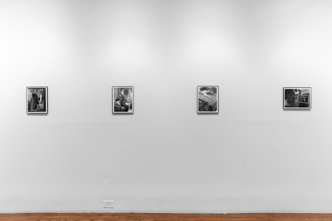 Installation view of four photographs in the exhibition. Four photographs are evenly hung on a white wall. The content is illegible due to the light's glare against the glass frame. The first three photographs are portrait while the last photograph is landscape. 