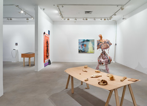 Large installation view that spans across two galleries. In the very back on the left, in the first gallery is a far away shot of a wooden table. Next to the table, further to the left, is a white pedestal with a black object on top of it. Above the pedestal is a painting that is illegible from the camera's distance from the object. Moving to the right and into the next gallery, sharing a wall back to back with the wooden table from the previous gallery is an orange piece of artwork. Above the work is a smaller canvas in a dark color and on the bottom of the orange is a purple stripe. On the middle wall in the gallery, is a painting of mostly blue, black, and light green colors organized in an abstract geometric form. In the front of these two works is an abstract sculpture in pink and orange. In front of this sculpture is a wooden table with various wooden objects placed around it. 