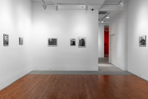 Installation view of six photographs hung on three white walls. There are two on the left-side wall, three on the middle wall, and one on the right-side wall. The images are illegible due to the camera lens and the glare of the frames. 