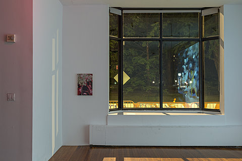 A small framed drawing is on a white wall next to a window. The reflection of a wall projection can be seen in the window.