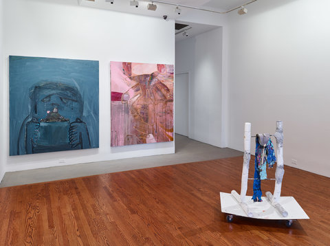 Installation view of the exhibition featuring a wall with two paintings side by side. On the left, the painting features an abstract figure outlined in black with a dark blue, teal background. On the right, is an abstract painting in pink with large outlines of what looks like hands. In front of the works on the floor, is a sculpture featuring white base on wheels. On top of the white, rectangular base are painted white tubes that lay horizontal while more tubes come out of the work vertically, stacking on top of one another. Rods connect the two poles of white tubes like a ladder. Draped over the rungs are items painted roughly in blue.   