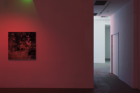 A square print hangs on a wall lit with red light next to a hallway leading to a white room.