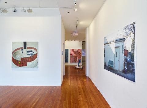 Installation view of the exhibition featuring two walls in a gallery and the hallway that leads into another gallery. On the left wall is a painting of what looks like a red fence in a circle with a swirling beige background. On the back of the fence-like structure is a black vertical rectangle resembling a doorway into the abyss. On the right wall, is a blown up photograph of a outside, backyard shed scene. A plastic chair leans up against a white building next to a door. On the other side of the structure is a grill, foliage, a cooler, and other mix of trash and objects outside on the cement. 
