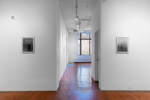 Installation view of two walls in the exhibition. The two walls are separated by a hallway leading to the front of the gallery. On the left wall there is a black and white photograph and on the right wall there is a black and white photograph, however, the glare of the camera lens and the glass make it the content less legible. 
