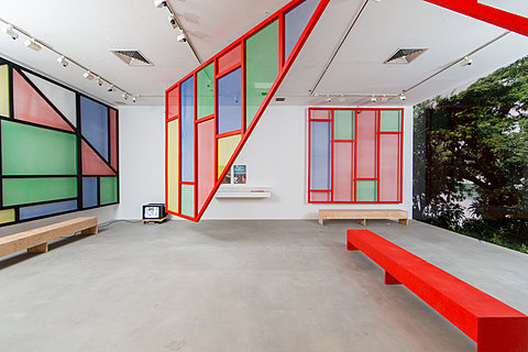 A gallery room with a red bench and a large, colorful geometric structure hanging from the ceiling. Several other geometric constructions are mounted on the walls.