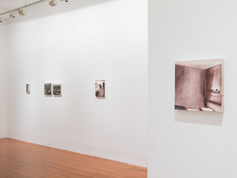 Installation image featuring two walls and five artworks in the exhibition. In the background and left most wall, four artworks are hung against the white wall. The farther ones are illegible from the cameras viewpoint. The closer image, on the left most side of the screen features a blurry purplish sketch in paint of a room. 