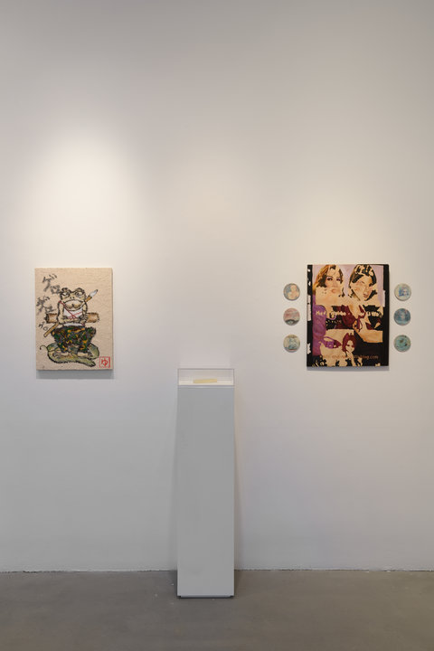 Installation view of three objects in the exhibition against a white wall. The first object is a painting with an anthropomorphic frog figure crossing its arms and wearing clothes as it balances a top a lily pad. Next to this frog, a white pedestal sits against the wall with a small glass box on top. The contents of the pedestal are illegible. On the right side of the pedestal, a painting hangs with three circular discs on each side. 
