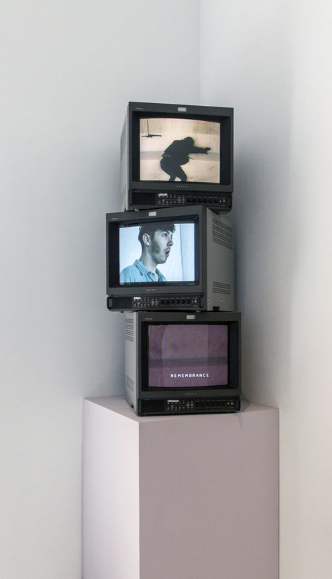 Installation view of three small television monitors stacked on top of one another sitting atop a white pedestal. The monitors are on and show various stills from the videos that play on them. On the top monitor, a figure is blurred and crouched away from the camera. In the middle, a figure's head and shoulders is shown as the figure looks off screen with a gaping mouth. The bottom monitor consists of a blurry static screen with the word "REMEMBRANCE" on the bottom in white type face. 