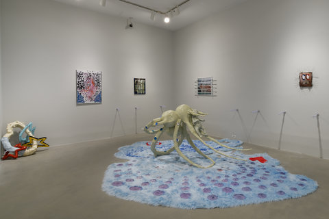 The gallery room pictured shows a large sculpture on the floor. The sculpture is of a spider-like form and is holding a baby in its pinchers. The sculpture is a cream color with blue reflective discs placed into its legs. The sculpture sits on a large blue and purple spotted fuzzy rug. On the walls behind the sculpture is a row of glass light-up rods coming off the wall. On the right wall above these rods there is a small painting with metal spikes hanging to the right, and a slightly larger painting barred off with metal rods hangs to the left. On the left wall there is a smaller hanging piece to the left and to the right there is a medium-sized abstract painting. In the left of the shot we see a cream rounded sculpture form with geometric colored shapes hanging from it that is propped against the wall. 