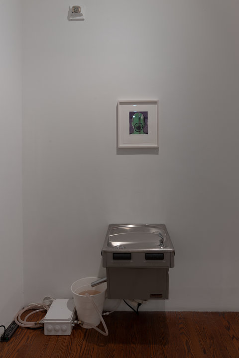 Installation image of a section of the exhibition featuring a water fountain mounted to the wall. Above the water fountain, hangs a picture in a white picture frame. 