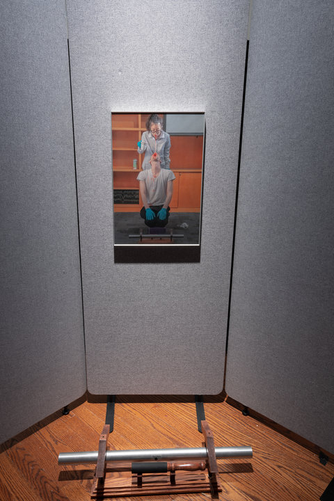 Close up image from a section of the installation featuring a photograph mounted onto a grey wall divider. The photograph features a figure in a white coat spitting red liquid into another figures mouth who looks up at the first figure. The second figure sits on their knees with their head tilted back with their mouth wide open. Both figures wear blue latex gloves. On the floor in front of the divider, a metal bar and wood contraption is placed on the floor. 