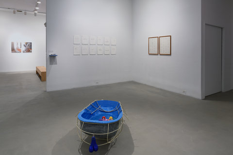 There is a kiddy pool-like sculpture in the middle of the room pictured. This form is lined with blue on the inside and has a grey exterior. There is also a wooden frame built around this sculpture. The inside of the sculpture is filled with water and has a ring of rubber duckies floating in it. On the two cornered walls behind the sculpture we see a series of twelve drawings on the left wall and two drawings on the right wall. Also, on the edge of the left wall there is. small blue shelf stacked with papers. In the left of the shot we see into the gallery behind these walls which has a wooden bench and two photographs hanging on the back wall. 