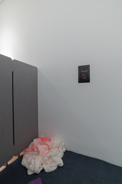 Installation view of a section of the exhibition featuring a small poster on the wall. A grey wall divider creates a small divide. Inside the divide is a carpet an a fluffy white blanket with pinkish red liquid sprayed on top of it. 