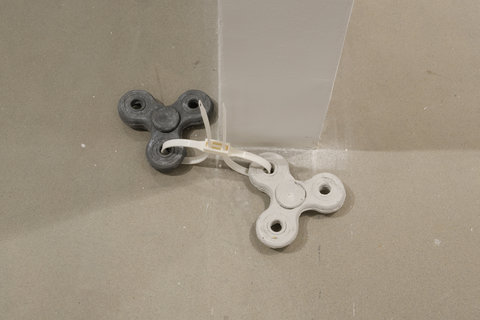 The image shows the floor beside a dividing wall that separates two galleries. On the floor laying beside the bottom of the dividing wall is two fidget spinner forms, one steel grey the other a grayish-white, looped together with a thick white zip-tie. 