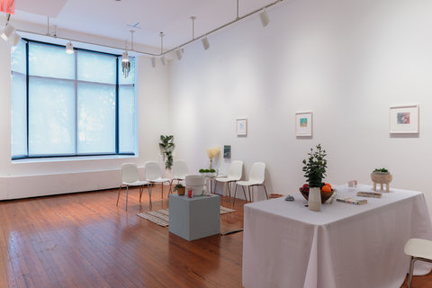 Installation view of an installation in the exhibition resembling a waiting room complete with a white table clothed, table, chairs, plants, fruits, and paintings on the wall. 