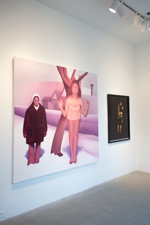 Installation view of two works in the exhibition hung on a white wall in front of a cement gallery floor. On the left, a painting is painted with pink tones and painted so there is a blurred effect over the entire canvas. The scene consists of two figures outside in the snow. On the left, the figure wears a white cap and a dark winter coat, while on the right, a figure is mostly nude. Behind the figures, a tree looms with a house in the background. On the right, the painting consists of a figure with various yellow containers balanced on their back along their spine. The entire image is a bit blurred and out of focus from the camera angle. 