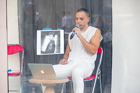 Viewed through a window, a person wearing all white sits in fronts of a laptop while speaking into a microphone. On the window is taped an image of a person.