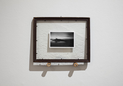 Image of a single work in the exhibition. A photograph is encapsulated by a wooden frame. The photograph in the wooden frame shows a landscape scene of a sky and lake view. 