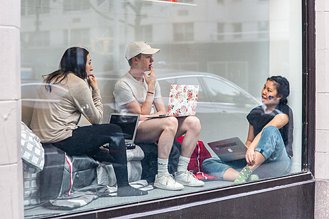 Viewed through a window, three people sit in a small window display. They all are holding laptops and are engaged in conversation.