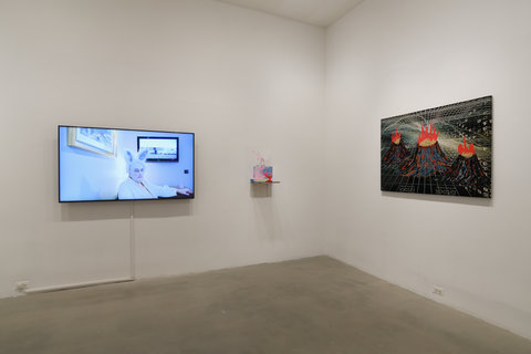 The image shows a corner of a room that has white walls and a concrete floor. On the right wall there is a woven textile piece stretched over a frame that features volcano-like shapes. Closest to the corner on the left wall is a small sculptural piece sitting on a platform coming off the wall. The sculpture is mostly pink, blue, and red. Next to the sculpture on the left wall is a TV with an image of a man dressed up as a bunny on it. 