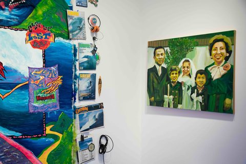 Image of a work in the exhibition featuring a very vibrant and multi colored abstract painting that is lined with vintage surfer magazines that have ocean motifs on the covers. Next to this work, on the right, is a painting of a family portrait, smiling into the camera. A green tint sets the mood of the painting. 