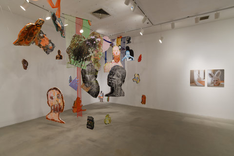 The image shows a room with a hanging installation of cut-out foam pieces and tule. On the cutout foam pieces are paintings and photographs. Subjects include portraits of people, cars, and abstract forms. On the right wall we see two medium-sized printed photographs of an older man dressed as a bunny. 