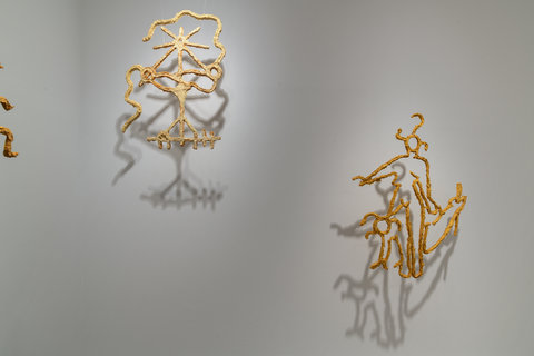 Installation image of two sculptures in the exhibition. The abstract sculptures of paper mache or cardboard material hang from the ceiling, just off the wall. The material swirls in organic and abstract shapes and the shadows from the objects reflect onto the back of the wall. 