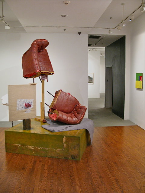 Installation view of the gallery. On a wood floor sits a platform. A couch is displayed, ripped in half, down the middle vertically. The top half of the couch is on a stilt. The bottom half lays below it on a folded textile. Also on the platform is a wood panel. Behind the structure is a cloud sculpture on a metal pole. 