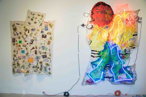 Installation image of two works in the exhibition. On the left, off-white sheets are hung on the wall in the shape of a pillow and blanket, plastered with various objects and cut out pieces of paper in various colors. Next, on the right, a mixture of tapestry and neon lights are arranged in a rainbow of colors and lit up with lights.  