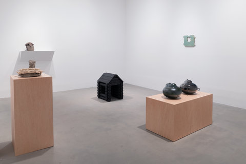 Installation image of five sculptures in the exhibition. In the center of the image, on the ground, sits a black log cabin. On the left, in the foreground, a ceramic and cement sculpture sits atop a light brown wood pedestal. In the left background, a stone draped with metal sits atop a protruding white shelf mounted on the wall. On the back wall, an enlarged, bread clip is mounted to the wall. It is light green. On the right of the image, two ceramic sculptures in dark glaze sit atop a light brown wooden bench pedestal. 