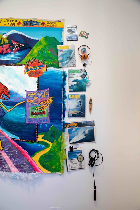 Image of a work in the exhibition featuring a very vibrant and multi colored abstract painting that is lined with vintage surfer magazines that have ocean motifs on the covers. 