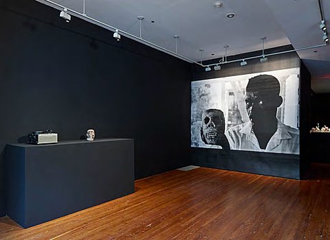 In a gallery with black walls is a large black-and-white projection of a person holding a ceramic skull.