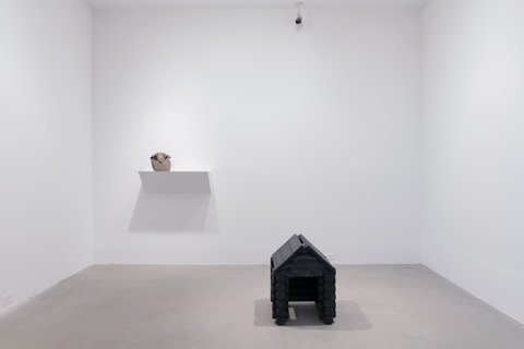 Installation image of two sculptures in the exhibition. In the foreground, on the grey cement ground, sits a small black, log cabin sculpture. Behind it, on the white wall, on a small protruding shelf, sits a stone with a piece of metal draped over it.  