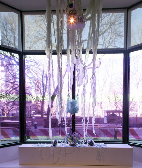 Installation view of the exhibition featuring four window panels. Several white, fabric streamers hang from the ceiling near a projector. A blue plastic bag also hangs from these strips of fabric. The windows are covered in a pinkish and purple film that changes the color of the light as it streams in. 