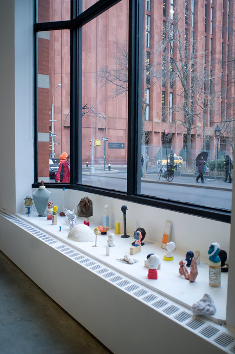 Installation view of objects seated on the window sill lit up from the light pouring in from the window panes. Several abstract clay and found-object works are scattered around the sill. 