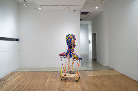 Installation view of the exhibition featuring an abstract sculpture in the center of the gallery space surrounded by white walls, sitting on top of a wooden floor. The sculpture itself consists of a wooden structure on wheels, wrapped with blue, orange, red, and yellow bands of string or tape.  