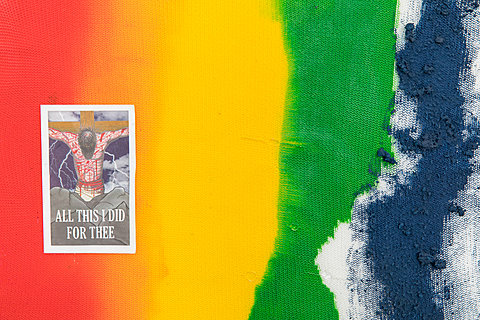 A detail of a large rainbow-painted mesh sheet, showing a card with the text "All this I did for thee," and a bloody, crucified figure.