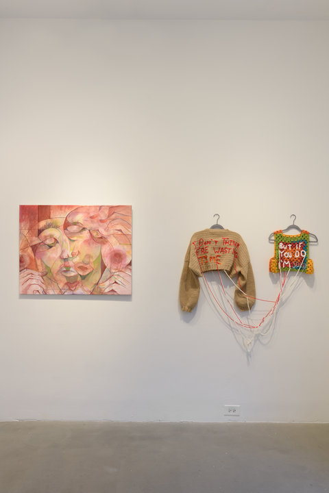 Installation view of two walls in the exhibition An abstract painting of faces in various pink pastels, hangs against a white wall. Right of this painting, are two hung pieces of clothing. On the right, a long-sleeve tank sweater with the words, “ I DON’T THINK WE’RE WASTING TIME” stitched in pink. On the right, a multicolored tank top is knitted with the words, “BUT IF YOU DO I’M SORRY” in white yarn. They are hung on the wall on grey hangers and attached to one another with long pieces of yarn. 